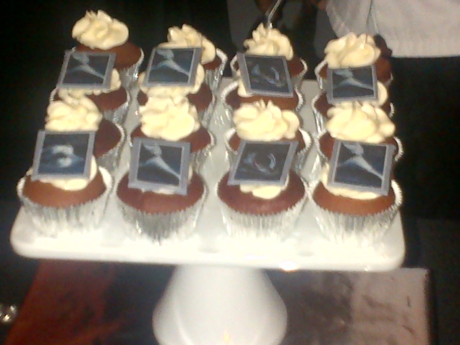 a tray with 12 vanilla cupcakes with white frosting in 3 rows of 4. The front and back rows have decorations in greyscale on them
