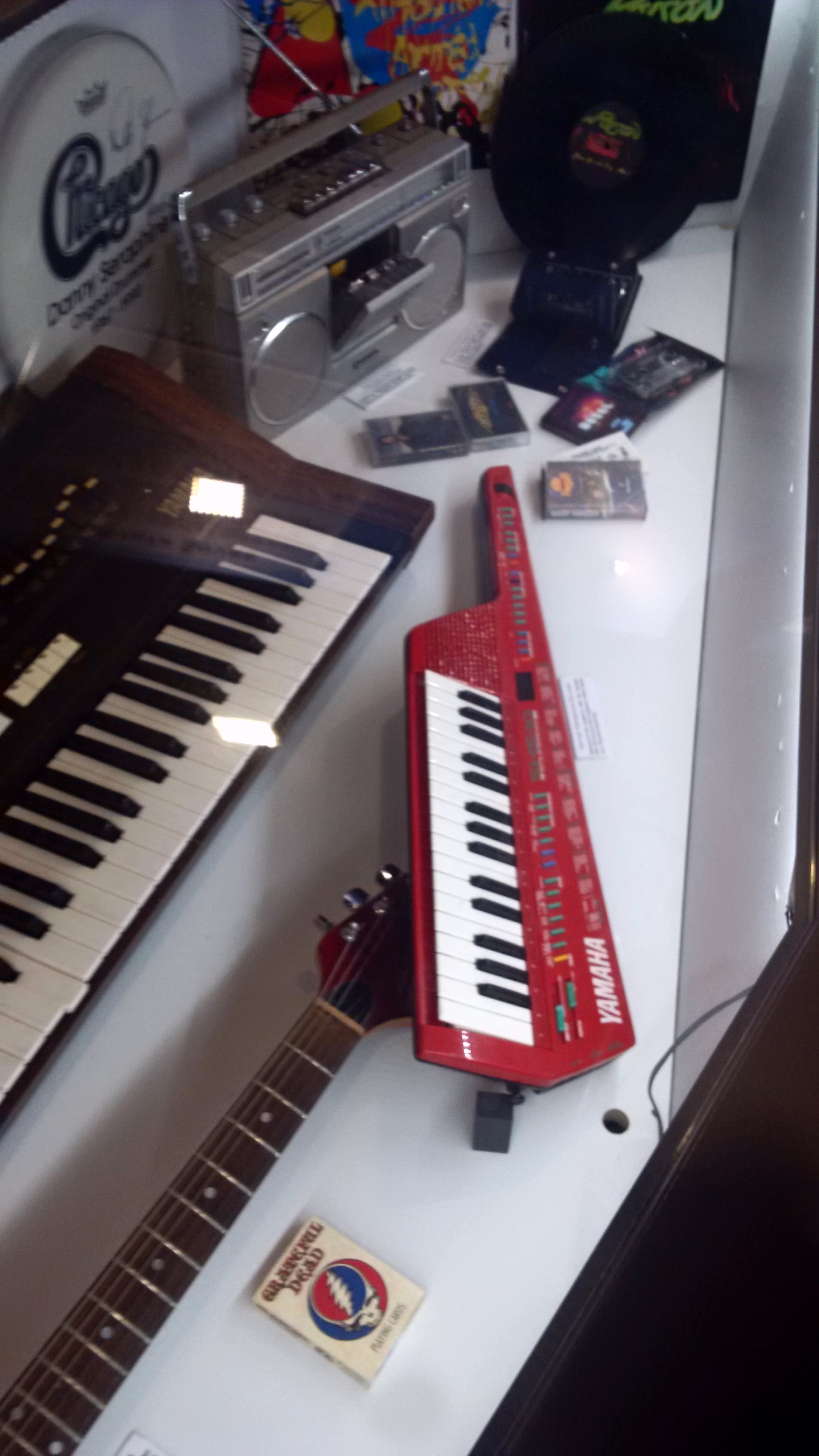 A collection of instruments and music memorabelia in a display case including a red keyboard with a guitar neck and a strap, a boom box portable stereo, cassette tapes, and an electronic keyboard.