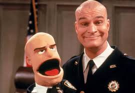 a bald man in a court officer's uniform holds up a puppet.