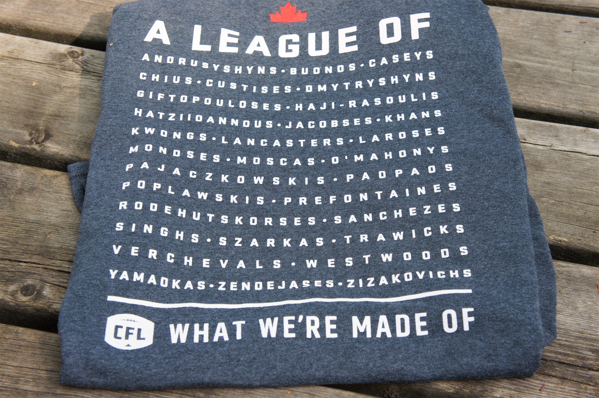 the back of the shirt - At the top it says a League of, followed by a list of multicultural names of former CFL players and at the bottom is the CFL logo and the words What We're Made Of